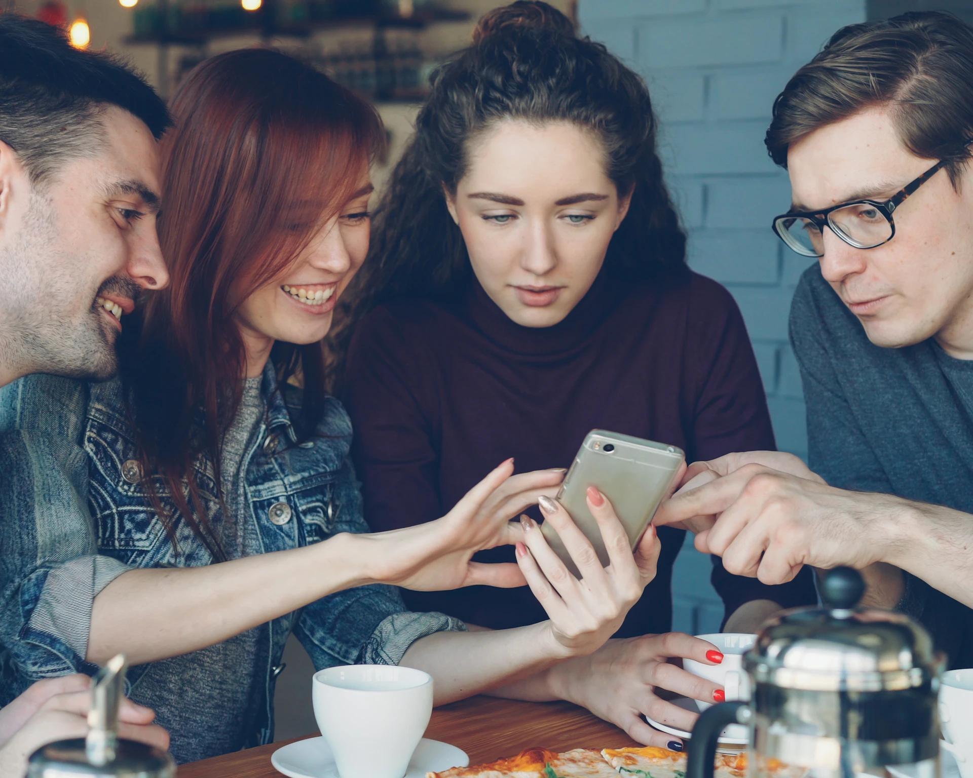Four friends laughing as they look at a phone together in a café