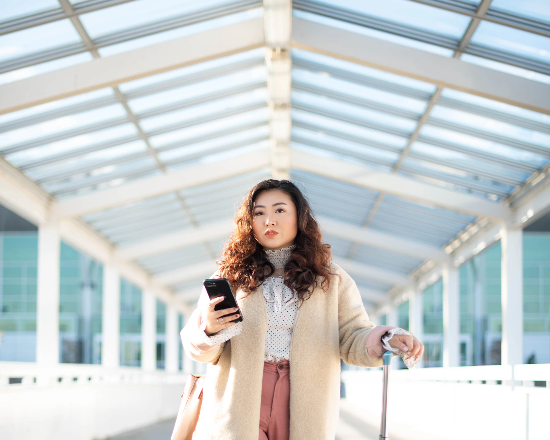 A woman holding her phone in a bright glass walkway, with a suitcase beside her