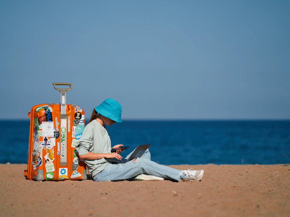 A person working on a laptop on a beach beside a suitcase