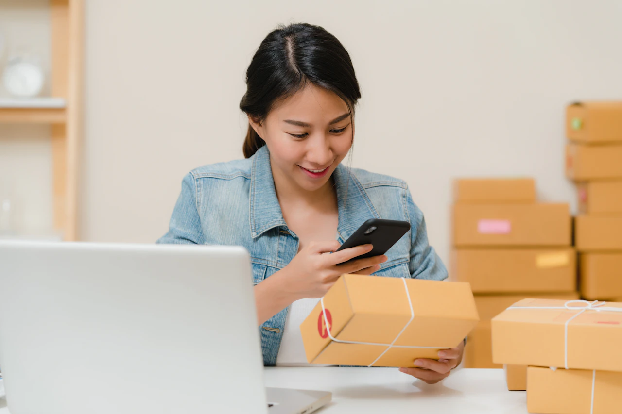A woman checking an online order on her phone beside a laptop and parcels